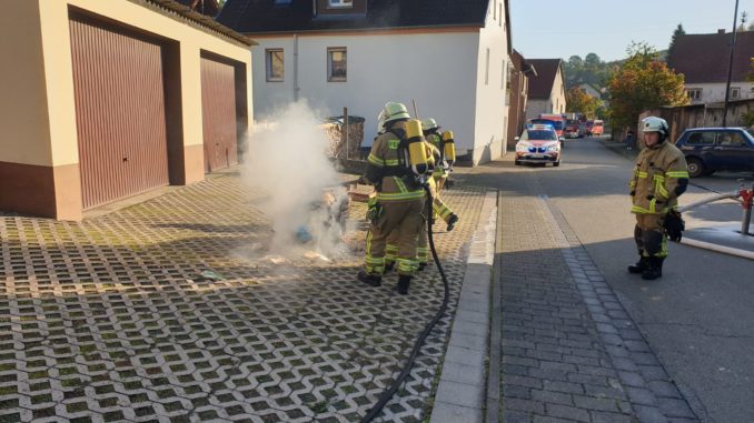 Mit Atemschutz ausgerüstete Einsatzkräfte löschen einen brennenden Gegenstand außerhalb des Gebäudes ab.