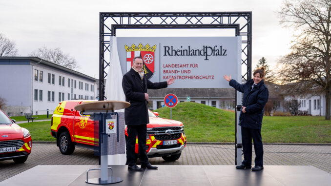 Innenminister Michael Ebling und Landesbrand- und Katastrophenschutzinspekteurin Kirstin Eisenhauer mit dem Schild der neuen Behörde. Im Hintergrund stehen landeseigene Einsatzfahrzeuge im neu gestalteten Design. Quelle: Innenministerium RLP