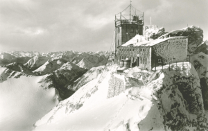 Die Wetterstation Zugspitze (Turm) mit dem direkt angrenzenden Münchner Haus vor 1938 (Herbst 1938: Sprengung des Westgipfels; hier im Hintergrund rechts) Quelle: Bildkunst-Verlag Gg. Poppe, Bad Kissingen / Aufnahme: H. Huber